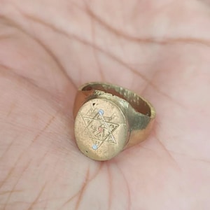 May include: A gold-toned metal ring featuring a flat, oval-shaped face. The face is engraved with a six-pointed star design containing a central symbol and two small white dots positioned at the top and bottom points of the star engraving.
