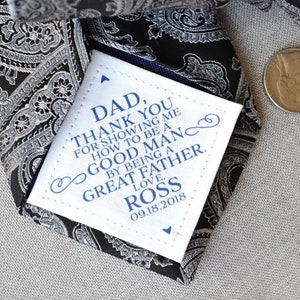 May include: A white tag with a blue border and text that reads "DAD, THANK YOU FOR SHOWING ME HOW TO BE A GOOD MAN BY BEING A GREAT FATHER. LOVE, ROSS 09.18.2018" attached to a black and silver paisley patterned tie.