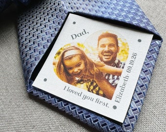 May include: A blue patterned tie with a personalized tag featuring a photo of a father and child. The tag reads "Dad, I loved you first. Elizabeth - 09.19.26."