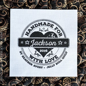 May include: A square white fabric label featuring a black circular stamp design with a central heart, the name Jackson, the year 2016, and the text Handmade for with love by Heather Avery Jelly Roll Quilt, centered on a black and gold patterned fabric.