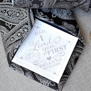 May include: A black and silver paisley patterned tie with a white tag attached. The tag has the text "Dad, I Loved you First" with a heart and the name "Brianna" and the date "09.19.2018".