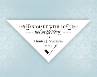 May include: White triangle label with black text that reads "Handmade with love - not perfection - by Christen J. Shaphanial 2024". A black needle and thread are drawn below the text.