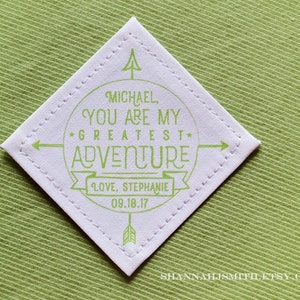 May include: A white diamond-shaped fabric patch with green stitching around the edges. The patch has a green circle with the text "MICHAEL, YOU ARE MY GREATEST ADVENTURE" and a banner with the text "LOVE, STEPHANIE 09.18.17".