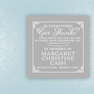 May include: A gray square plaque with white text. The text reads "In Everything Give Thanks" and "In Memory of Margaret Christine Cash, August 5, 1976 - March 1, 2015." Decorative flourishes frame the text. The background is a light blue floral pattern.