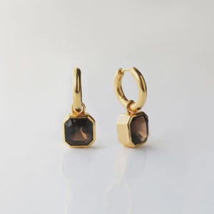 May include: A pair of gold-colored hoop earrings with a square, dark brown gemstone pendant. The earrings are displayed on a white surface, showcasing their polished finish and geometric design. The gemstones have a faceted cut.