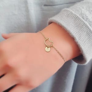 May include: Gold-colored bracelet featuring a cat silhouette charm and a small circular charm with the letter "N". The bracelet is delicate and appears to be made of a thin chain. The bracelet is worn on a wrist.
