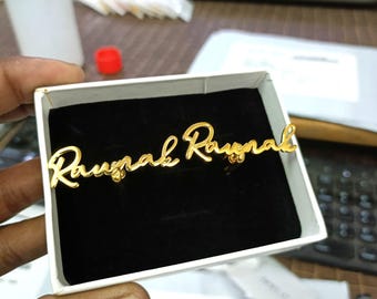 Puede incluir: Un pin con el nombre dorado, con la inscripción "Raunak Raunak" en cursiva, se muestra dentro de una caja blanca con interior de terciopelo negro. Un accesorio personalizado.