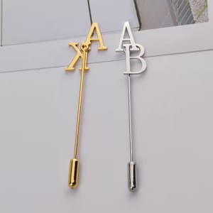 Puede incluir: Dos prendedores, uno dorado y otro plateado, cada uno con una letra mayúscula. El prendedor dorado muestra las letras "KA", mientras que el plateado muestra "AB". Ambos prendedores tienen una aguja larga y delgada.