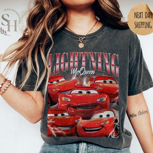 Puede incluir: Camiseta gris oscuro con un dise&ntilde;o de Rayo McQueen de la pel&iacute;cula Cars. El dise&ntilde;o incluye m&uacute;ltiples im&aacute;genes del coche de carreras rojo con el texto "LIGHTNING McQueen" en rojo y blanco. La camiseta es de color pimienta.