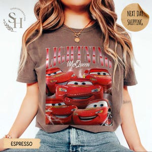 Puede incluir: Camiseta color espresso con un dise&ntilde;o de Rayo McQueen de la pel&iacute;cula Cars. El dise&ntilde;o incluye el texto "LIGHTNING McQueen" en rojo y blanco, con m&uacute;ltiples im&aacute;genes del coche de carreras rojo. La camiseta tiene un aspecto vintage y desgastado.