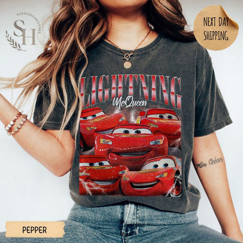 Puede incluir: Camiseta gris oscuro con un dise&ntilde;o de Rayo McQueen de la pel&iacute;cula Cars. El dise&ntilde;o incluye m&uacute;ltiples im&aacute;genes del coche de carreras rojo con el texto "LIGHTNING McQueen" en rojo y blanco. La camiseta es de color pimienta.