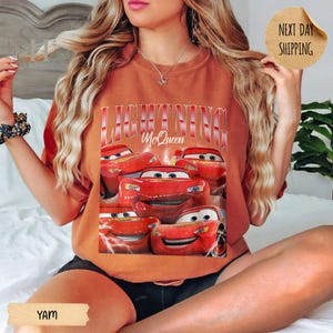 Puede incluir: Camiseta de color &oacute;xido con un gr&aacute;fico de Rayo McQueen de la pel&iacute;cula Cars. El dise&ntilde;o incluye m&uacute;ltiples im&aacute;genes del coche de carreras rojo y el texto "LIGHTNING McQueen" en una fuente estilizada. La camiseta tiene un corte relajado.