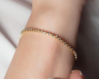 May include: A gold-colored bracelet with small, clear gemstones. The bracelet is worn on a wrist, and the background is a white shirt.