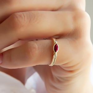 Puede incluir: Un anillo de oro con una piedra preciosa roja oscura con corte marquesa. El anillo tiene una banda texturizada y está adornado con pequeñas piedras preciosas redondas y claras a ambos lados de la piedra central. El anillo se muestra en un dedo.
