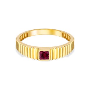 Può includere: Un anello d'oro con una fascia a coste e una gemma quadrata, rosso scuro. La superficie lucida dell'anello riflette la luce. La gemma è un punto focale. L'anello è posto su uno sfondo bianco.