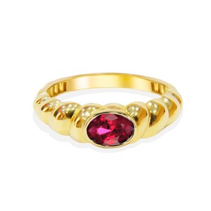 May include: A gold ring with a twisted band design and a vibrant oval-cut red gemstone. The ring is set against a white background, highlighting the polished gold and the gemstone's rich color.