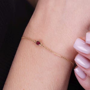 May include: A delicate gold chain bracelet featuring a single, small, oval-shaped deep red gemstone set in a simple gold bezel. The bracelet is thin and minimalist in its construction, showcasing a refined link design that encircles the wrist.