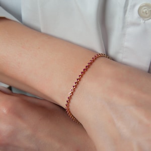 Könnte beinhalten: Ein zartes goldenes Armband, das mit kleinen, leuchtend roten Edelsteinen verziert ist. Das Armband ist dünn und flexibel und schmiegt sich angenehm um das Handgelenk. Die Edelsteine sind gleichmäßig verteilt.