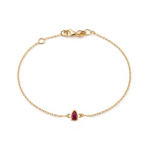 May include: A delicate gold chain bracelet featuring a single teardrop-shaped red gemstone set in a gold bezel. The bracelet includes a small circular link chain and a secure lobster claw clasp mechanism at the top for fastening the jewelry piece.