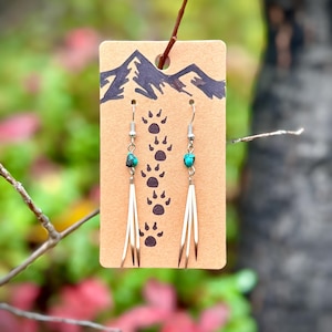 Puede incluir: Pendientes con cuentas turquesas y detalles de plumas cuelgan de ganchos plateados. Los pendientes se exhiben en una tarjeta marrón con una ilustración de montaña y un diseño de huellas de patas. El fondo es borroso.
