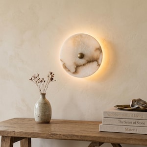 May include: A circular wall light with a marble effect, brown markings, and a brass fitting. A small vase with dried flowers is on a wooden table, with books and a decorative tray. The light is backlit.