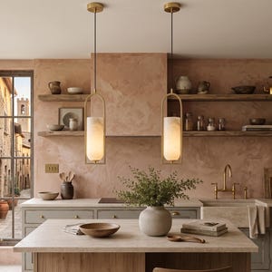 Op de afbeelding: Twee cilindrische hanglampen met messing frames en matglazen kappen hangen boven een kookeiland. De keuken heeft een stenen aanrechtblad, grijze kasten, open houten planken met diverse keramische voorwerpen en een getextureerde roze muur.