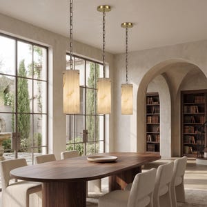May include: Three pendant lights hang above a wooden dining table. Each light has a cylindrical, off-white shade with vertical lines and a gold-coloured ceiling mount. The lights are suspended by chains. The room has large windows and a neutral colour palette.