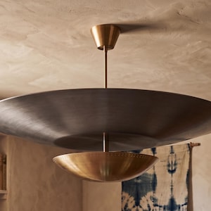 May include: A ceiling-mounted pendant light fixture featuring a large, flat, dark metallic disc suspended by a central rod, positioned above a smaller, bowl-shaped brass component with a perforated edge detail, creating a layered, dual-tone design.