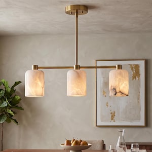 May include: A three-light linear chandelier featuring a brushed brass finish metal frame and three cylindrical, translucent white glass shades with subtle grey marble-like veining patterns, suspended from a matching brass ceiling canopy.