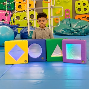 May include: A young person stands behind four colorful, illuminated geometric shapes: a yellow square with a diamond, a blue square with a circle, a green square with a triangle, and a purple square with a mirrored interior. The shapes are on a blue mat.