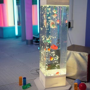 May include: A tall, rectangular aquarium lamp with colorful, animated fish. The lamp has a white base and top, with a clear, water-filled column. Bubbles and various toy fish float inside. Additional colorful cylinders and spheres are in the foreground.