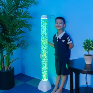 May include: A tall, cylindrical bubble tube with a white base and top, filled with green-lit water and floating bubbles. The tube has small, yellow fish decorations. The product is designed for sensory stimulation and is suitable for children.