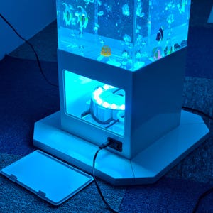 May include: A white, rectangular aquarium with a blue LED light. The aquarium has a clear, rectangular tank filled with water and decorative fish. The base has a rectangular opening with a light and a power cord. A detached white cover is in the foreground.