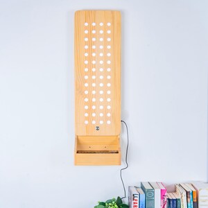 May include: A tall, rectangular light-colored wooden wall-mounted display board featuring a vertical grid of circular cutouts. A small shelf is attached to the bottom, and a thin black electrical cord extends from the side of the wooden structure.