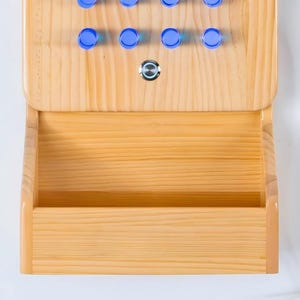 May include: A light-colored wooden wall-mounted organizer featuring a top section with eight circular blue pegs and a single central silver push-button switch, positioned above a wide, open storage compartment with a smooth, natural wood grain finish.