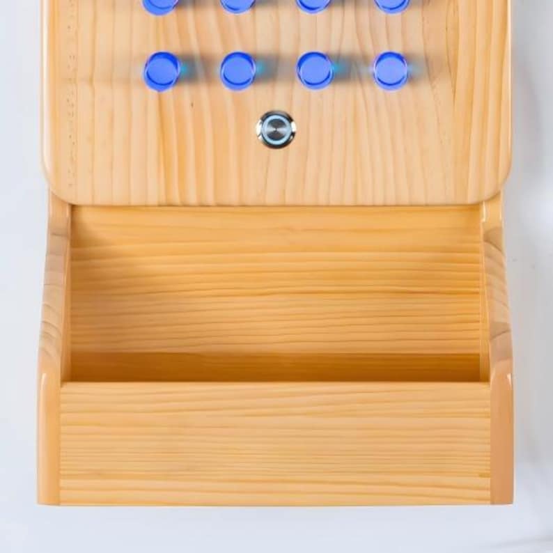 May include: A light-colored wooden wall-mounted organizer featuring a top section with eight circular blue pegs and a single central silver push-button switch, positioned above a wide, open storage compartment with a smooth, natural wood grain finish.