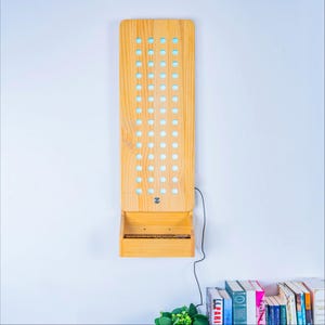 May include: A vertical light-colored wooden wall-mounted display board featuring a grid of circular cutouts and a small integrated shelf at the bottom. The board has a light blue backing visible through the holes and a black power cord extending from the side.