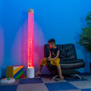 May include: A tall, illuminated bubble tube lamp with a white base and top, filled with red-tinted water and floating bubbles. A child sits near the lamp, with a black chair and a colorful cube toy nearby. The background is blue.