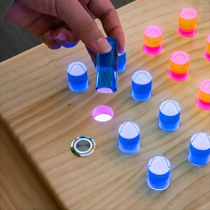 May include: A light-colored wooden board featuring multiple circular holes and a series of translucent, cylindrical pegs in shades of blue, pink, and orange. One blue peg is being placed into a hole, while a circular metal button is embedded.