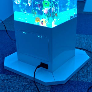 May include: A white, illuminated aquarium with colorful, cartoon-like fish floating inside. The aquarium has a rectangular base and a matching white cabinet with a door. A black power cord is plugged into the side. The aquarium is on a blue carpet.