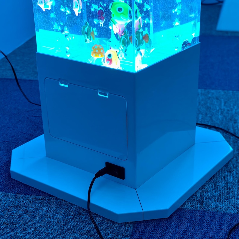 May include: A white, illuminated aquarium with colorful, cartoon-like fish floating inside. The aquarium has a rectangular base and a matching white cabinet with a door. A black power cord is plugged into the side. The aquarium is on a blue carpet.