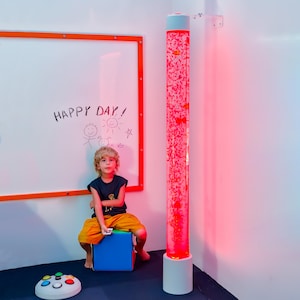 May include: A child sits near a tall, illuminated bubble tube with red and orange accents. The tube is mounted on a white base. A whiteboard with the words "HAPPY DAY!" and drawings is in the background. A game controller is on the floor.