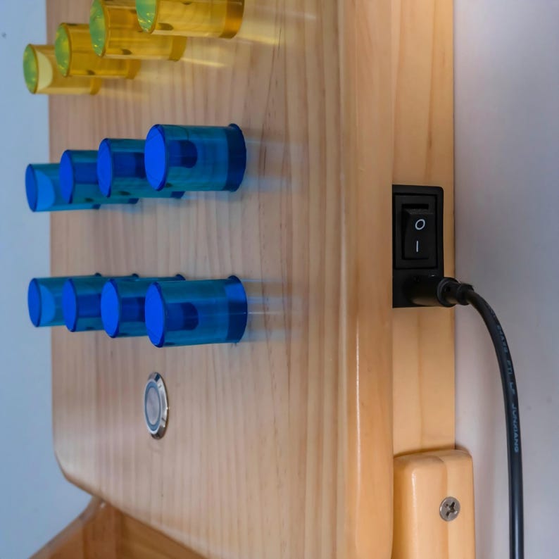 May include: A light-colored wooden panel featuring rows of translucent yellow and blue cylindrical knobs. A black rocker switch is mounted on the side, connected to a black power cable. A small circular metal button is embedded in the wood surface.