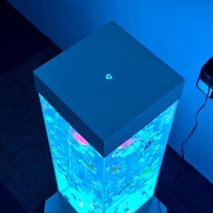 May include: A tall, illuminated aquarium with a square top and base. The tank is filled with colorful, artificial aquatic plants and fish, lit by a vibrant blue light. The top is a solid, light blue color with a small, illuminated symbol.
