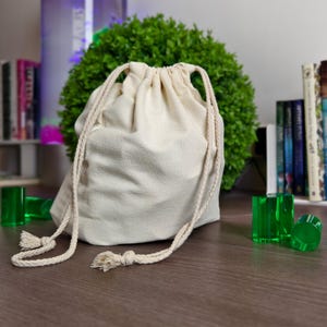 May include: A beige drawstring bag with a cream-colored cord and tassels. The bag is on a brown surface, with several green, transparent, cylindrical objects nearby. A green bush and books are in the background.