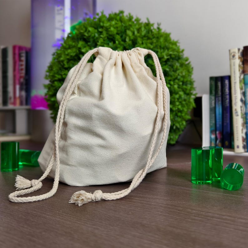 May include: A beige drawstring bag with a cream-colored cord and tassels. The bag is on a brown surface, with several green, transparent, cylindrical objects nearby. A green bush and books are in the background.