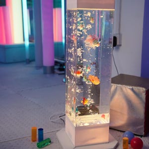 May include: A rectangular, illuminated bubble tube with colorful fish inside. The clear acrylic tube sits on a white base and is surrounded by colorful, cylindrical lights. Small toys are on the floor.