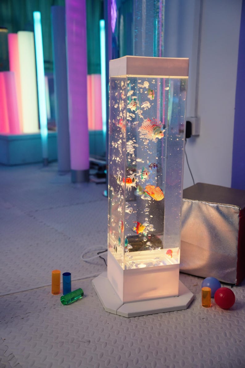 May include: A rectangular, illuminated bubble tube with colorful fish inside. The clear acrylic tube sits on a white base and is surrounded by colorful, cylindrical lights. Small toys are on the floor.