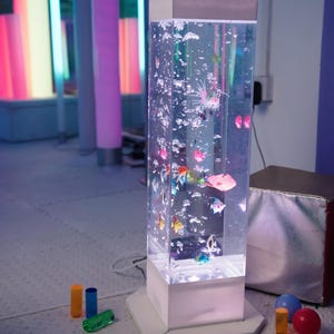 May include: A clear, rectangular bubble tube with colorful, plastic fish and bubbles. The tube sits on a white base and is surrounded by colorful, illuminated columns. Small, colorful toys are scattered on the floor.