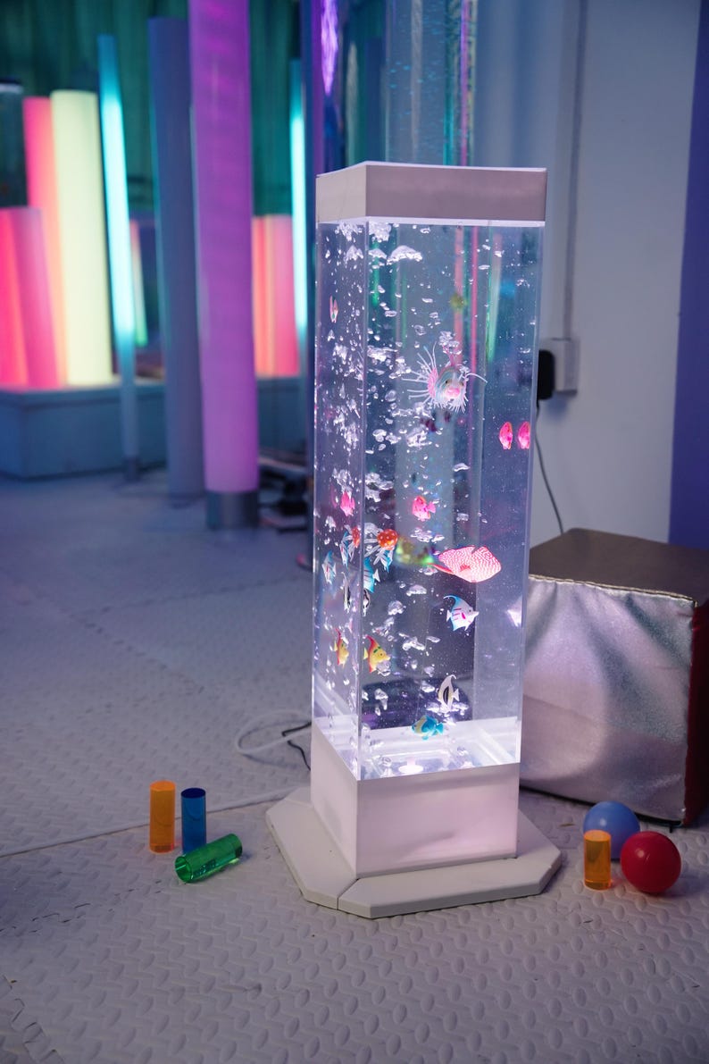 May include: A clear, rectangular bubble tube with colorful, plastic fish and bubbles. The tube sits on a white base and is surrounded by colorful, illuminated columns. Small, colorful toys are scattered on the floor.